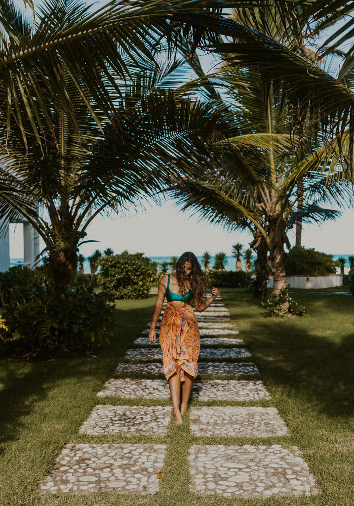 Woman wearing Kaiilani Fiji Blockprint sarong in orange floral pattern, standing on a path between palm trees with the ocean in the background.