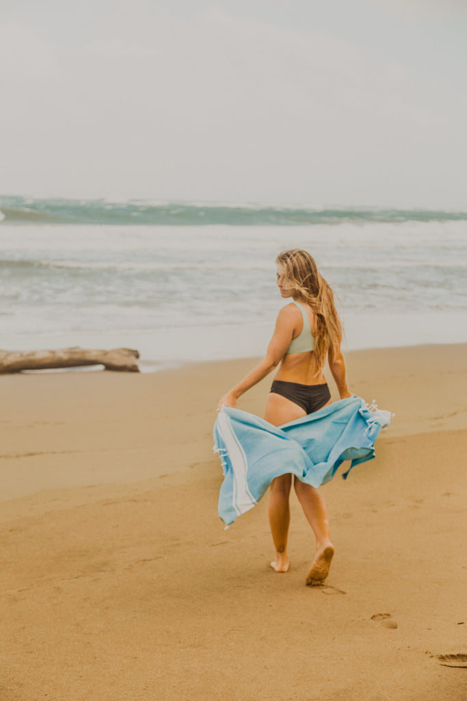 Woman walking towards the ocean, looking back with Kaiilani Gaia hand loom towel in Brisa, sky blue and white flowing behind her.