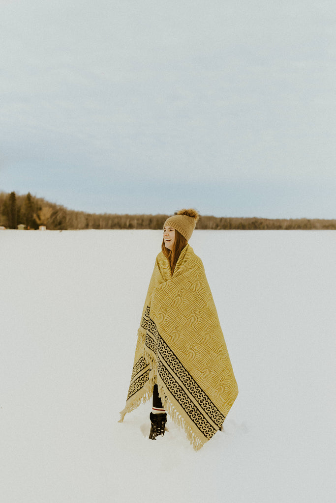 Kaiilani Kos Throw Blanket in Turmeric wrapped around woman's shoulders laughing in the snow. The Kos Throw blanket in turmeric features a swirling pattern in shades of gold with a black geometric border. Finished with fringe.