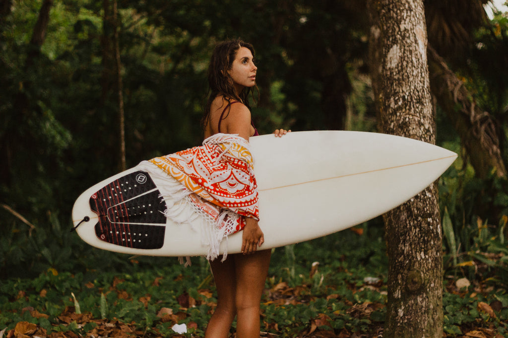 Women with a surfboard in the forest with a Kaiilani beach blanket mandala draped over her shoulders.