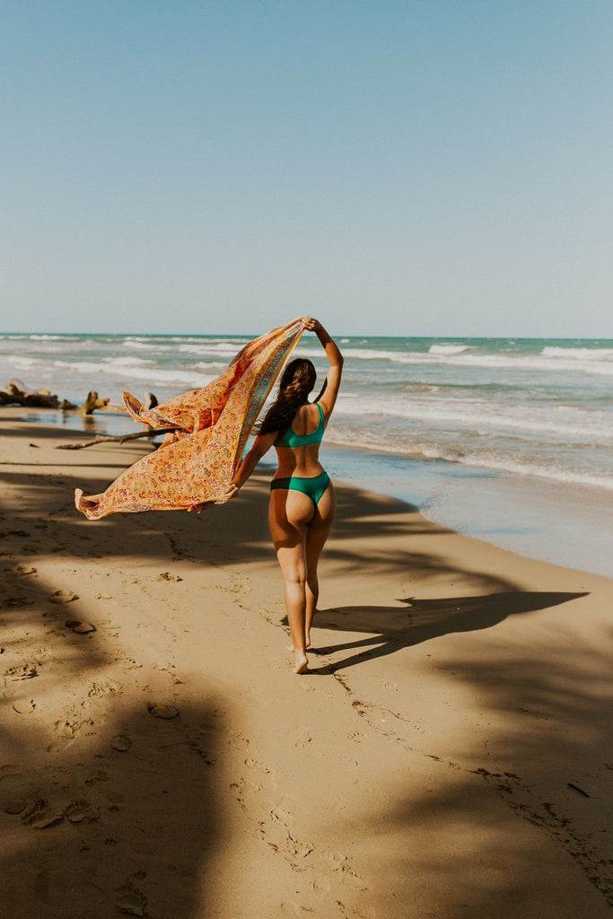 Woman holding Kaiilani Fiji Blockprint sarong in orange floral pattern, flowing in the wind on the beach.
