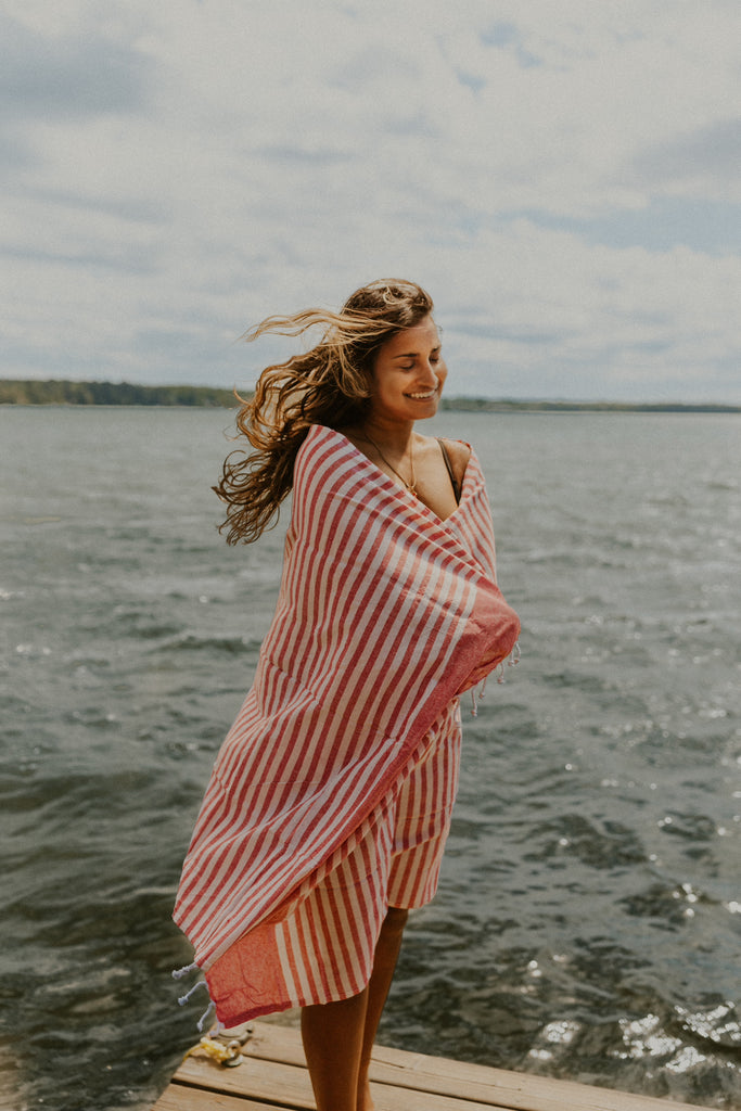 Karina standing on a dock with a lake in the background, wrapped in the Fuego towel, which is red and white striped with a hand tied fringe.