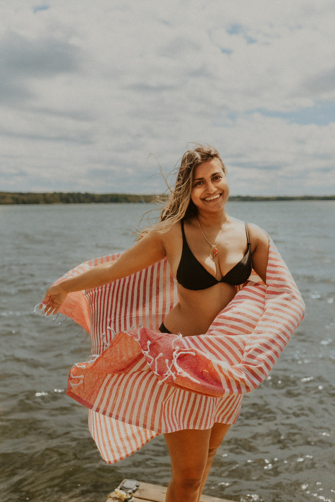 Karina smiling at the camera, standing on a dock with a lake in the background, wrapped in the Fuego towel, which is red and white striped with a hand tied fringe.