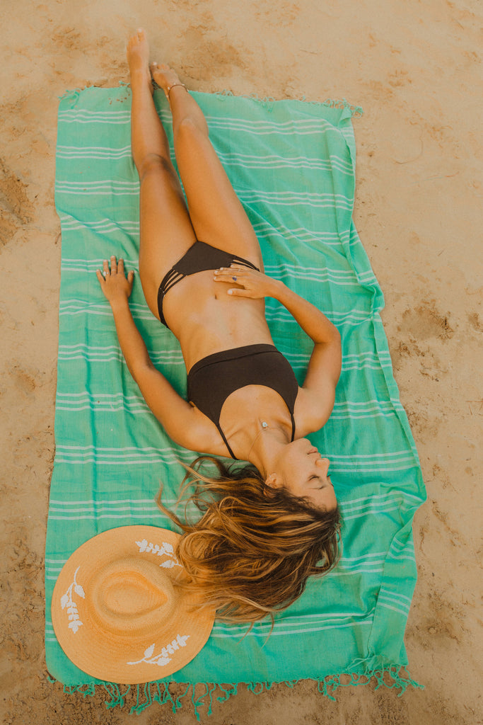 Woman lying down on Kaiilani Mélon turquoise hand loom towel on the beach.
