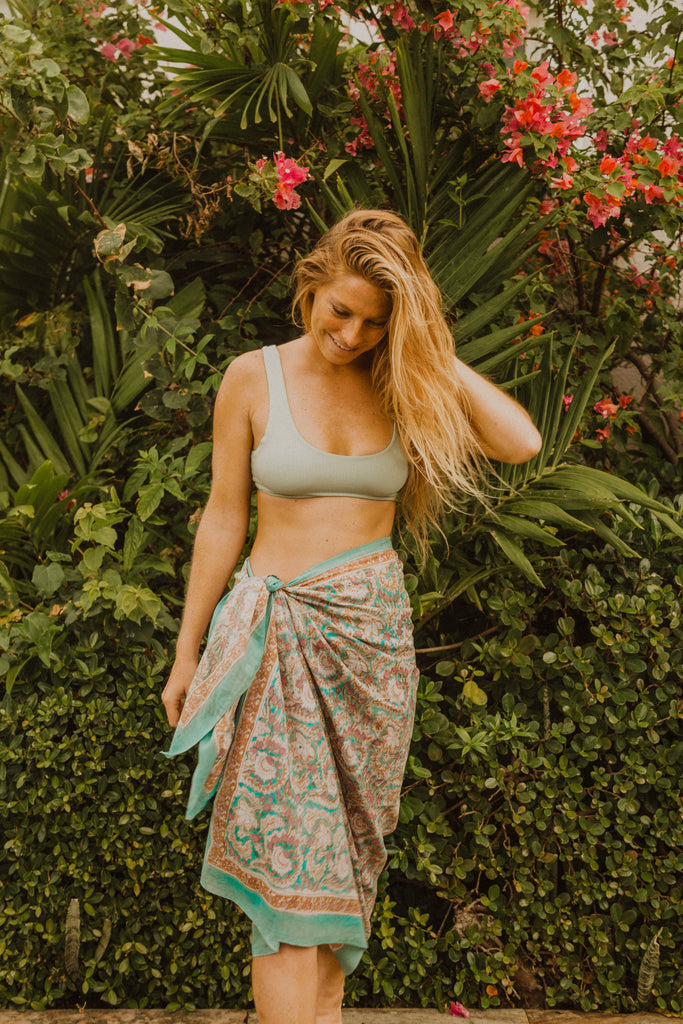 Woman standing in front of large jungle plants wearing Kaiilani Maldives blockprint sarong with aqua, lilac and beige floral pattern on it.