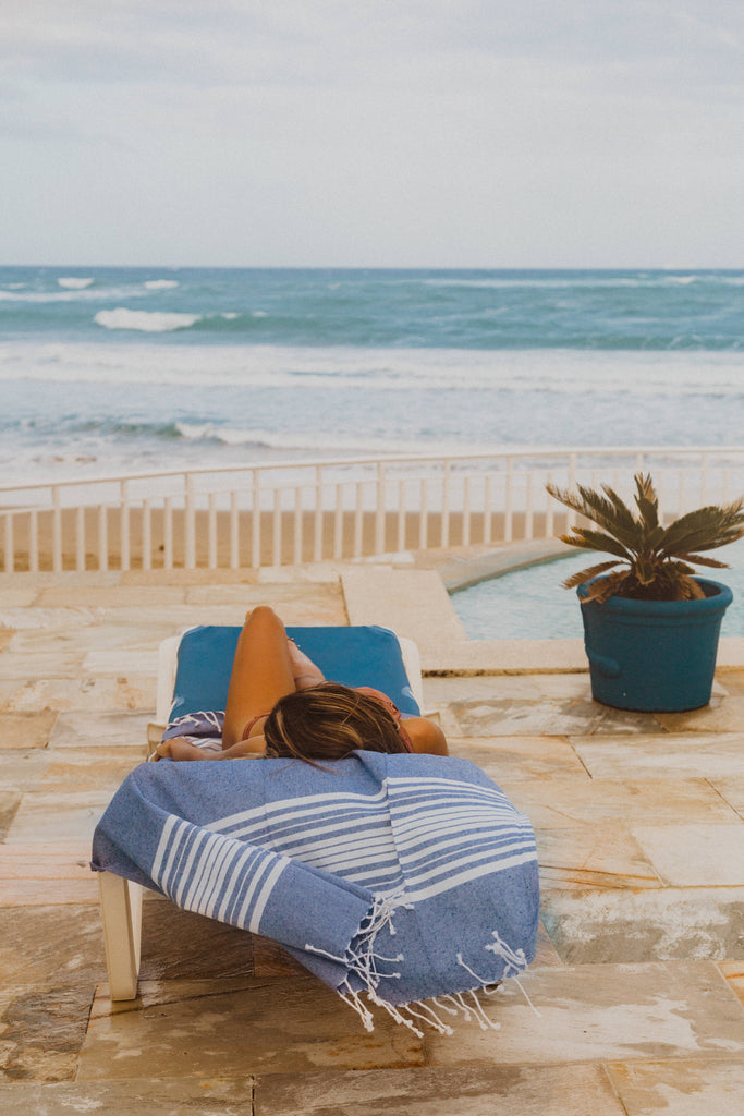 Woman on pool lounger facing the ocean lying on Kaiilani Oceano indigo and white hand loom towel.