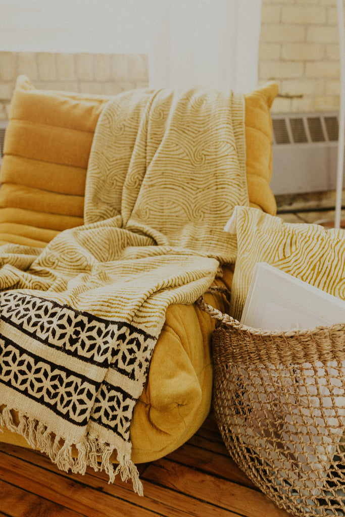 Kaiilani Kos Throw Blanket in Turmeric, thrown on a mustard floor chair.  The Kos Throw blanket in turmeric features a swirling pattern in shades of gold with a black geometric border.  Finished with fringe.