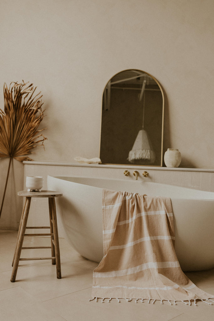 Kaiilani Gaia handloom towel in Arena, a sand colour, hanging off the edge of a curved free standing bathtub with mirror and dried florals in the back.