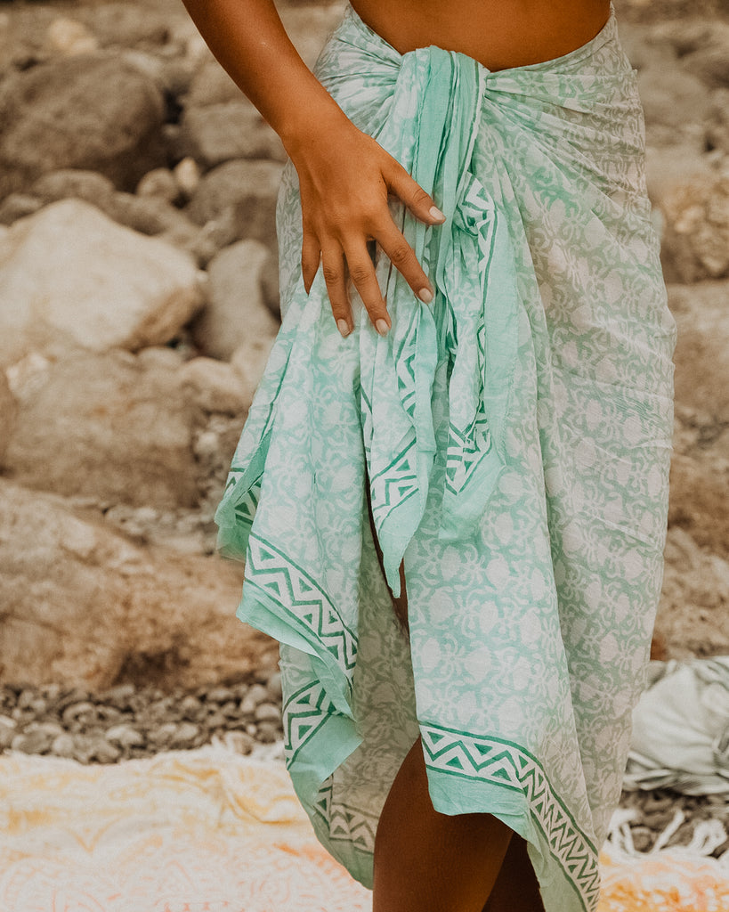 Women on rock on the beach wearing Kaiilani Positano Blockprint sarong with geometric pattern in aqua and white.